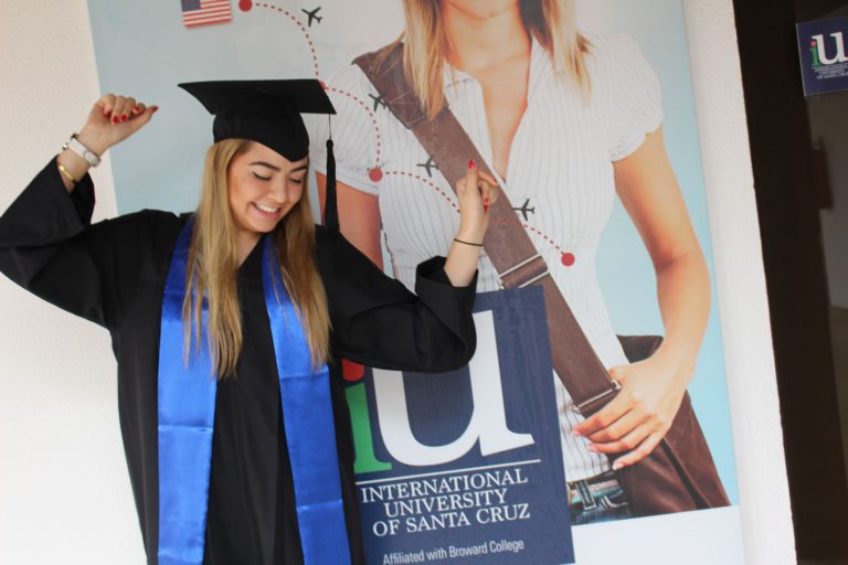 Graduada de iU con toga y birrete junto a un banner de International University of Santa Cruz